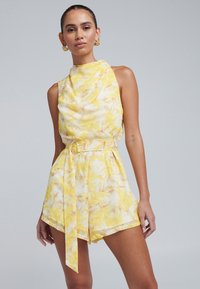 Geel-witte bloemenprint romper met een hoge hals, overslagstijl en bijpassende riem. Gladde stof, korte lengte en zijzakken.