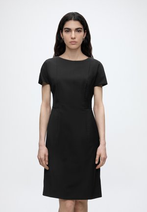 Žena s dlhými tmavými vlasmi, oblečená v priliehavých čiernych šatách s krátkym rukávom, stojaca pred jednoduchým svetlým pozadím, čelom vpredu.