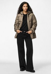 Giacca imbottita con cappuccio in taupe, caratterizzata da un design trapuntato e polsini elasticizzati. Abbinata a jeans neri a gamba larga e scarpe nere.