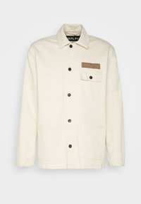 Chaqueta de color beige claro hecha de algodón, con cierre de botones en la parte delantera, dos bolsillos de parche en la parte inferior y un bolsillo en el pecho con una etiqueta de logo.