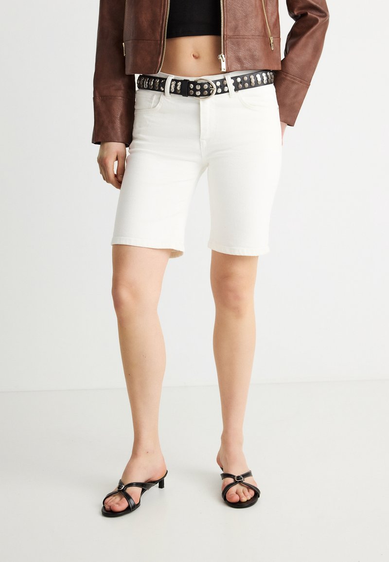 Γυναίκα που φοράει λευκά σορτς μέχρι το γόνατο, μαύρη ζώνη με καρφιά, καφέ δερμάτινο μπουφάν και μαύρα πέδιλα με τακούνι και ανοιχτά δάχτυλα, στέκεται σε λευκό πάτωμα.