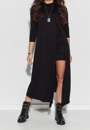 Zwarte lange mouw cardigan met een voorste split, gedragen over een aansluitende zwarte jurk. Gestructureerde stof en geaccentueerd met zwarte combatlaarzen met studs.