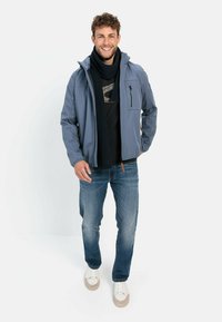 Lyseblå vandtæt jakke med en lynlåslomme, sort sweatshirt under, mørke jeans og hvide sneakers, suppleret af et mørkt tørklæde.
