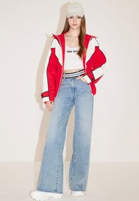 Chaqueta roja y blanca con cremallera, top blanco corto, jeans de pierna ancha azules y zapatillas blancas gruesas, complementados con una gorra gris clara.