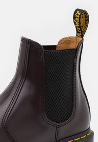 Botín Chelsea de cuero marrón oscuro con panel lateral elástico negro, forro interior color marrón claro, suela negra, costura amarilla y lengüeta con texto amarillo.