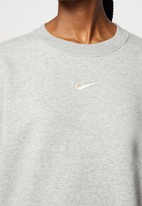 Detailní záběr na osobu oblečenou v světle šedém mikině s kulatým výstřihem, na jejíž hrudi je malé bílé logo Nike swoosh.