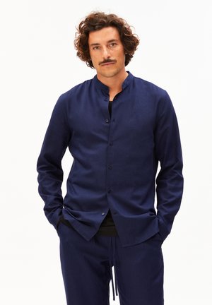 Mann mit lockigem Haar und Schnurrbart, trägt ein dunkelblaues Hemd mit Stehkragen und passenden Hosen, Hände in den Taschen, neutraler Hintergrund.