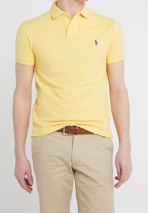 Man draagt een geel poloshirt met korte mouwen en een klein logo, gecombineerd met beige korte broek en een bruine gevlochten riem.