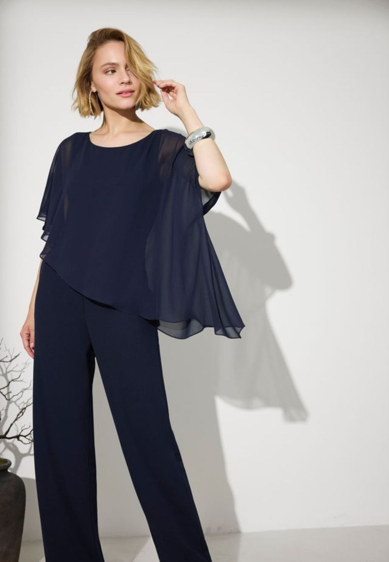 Kvinde med kort blondt hår iført flydende navyblå top og bukser med vide ben, rører ved håret og står ved siden af en bar gren i en vase mod en hvid væg.