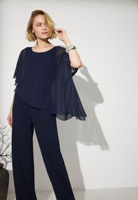 Femme aux cheveux blonds courts portant un haut bleu marine fluide et un pantalon à jambes larges, touchant ses cheveux, debout à côté d'une branche nue dans un vase contre un mur blanc.