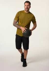 Olivgrön träningsskjorta med korta ärmar, matchad med svarta shorts, som håller en svart medicinboll, mot en neutral bakgrund.