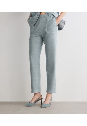 Vrouw draagt lichtgrijze nauwsluitende broek met steekzakken vooraan en bijpassende geweven slingback-hakken, staand tegen een effen muur.