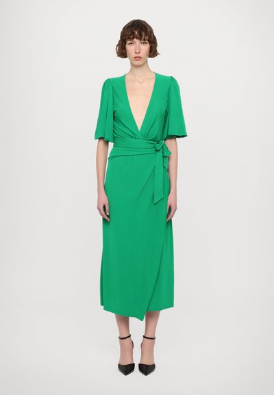 Green wrap dress with short, flared sleeves, deep V-neckline, and a side tie. The design features a mid-calf length and smooth texture. Black pointed heels.