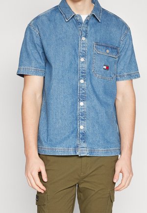 Mann trägt kurzärmeliges Jeanshemd mit Brusttasche und olivgrüner Cargohose, steht vor neutralem Hintergrund.