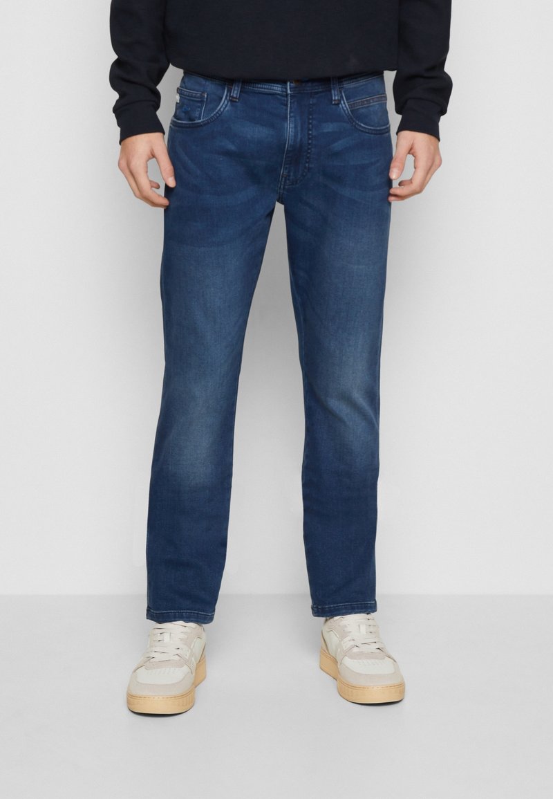 Homme portant un jean bleu foncé à jambes droites et des baskets blanches, debout devant un fond gris clair uni.