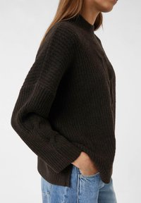 Maglione a coste marrone scuro con collo alto e maniche larghe, caratterizzato da un motivo a trecce lungo le braccia. Indossato con jeans azzurri chiari.