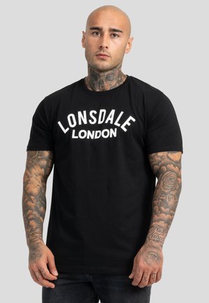 Skaldet mand med nakke- og armtatoveringer iført en sort Lonsdale London T-shirt, stående foran en ensfarvet grå baggrund.