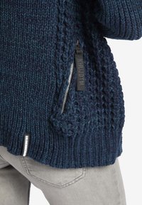 Pletený svetr v tmavě modrých odstínech s žebrovanou strukturou, vybavený boční kapsou na zip a logovou visačkou na lemu.
