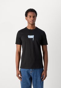 Camiseta de algodón negra con un pequeño gráfico azul en el pecho. Manga corta y cuello redondo. Combinada con unos vaqueros azules de corte holgado.