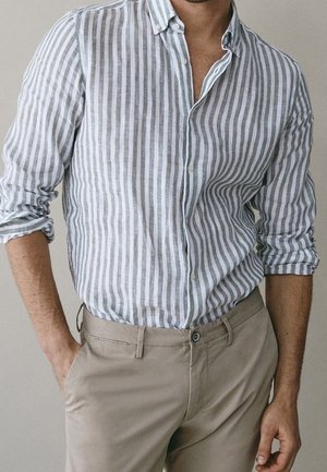 Mann trägt ein weißes und graues, vertikal gestreiftes Hemd mit hochgekrempelten Ärmeln und beigen Hosen, mit einer Hand in der Tasche.