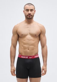 Chiloți boxer negri cu o bandă elastică roșie în talie, pe care este imprimat vizibil logo-ul Nike. Țesătura pare netedă și aderentă.