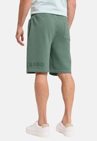 Grüne Baumwollshorts mit seitlichen Taschen, elastischem Bund und Druckdetail am linken Bein. Getragen mit weißen Sneakers.