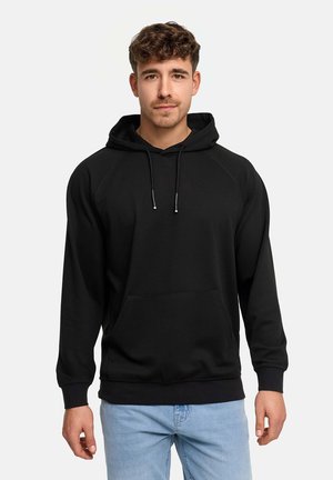 Joven con cabello corto y rizado que lleva una sudadera negra con capucha y vaqueros azul claro, de pie frente a un fondo liso.