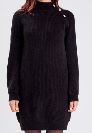 Robe pull noire en maille côtelée avec col montant et trois boutons argentés sur l'épaule, portée par une personne aux longs cheveux.