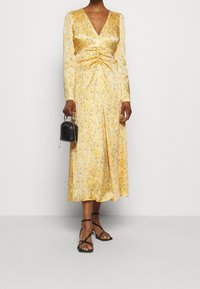 Gul blommönstrad midikjol med strukturerade långärmar, en djup urringning och samlad detalj vid midjan, kombinerad med svarta sandaler.