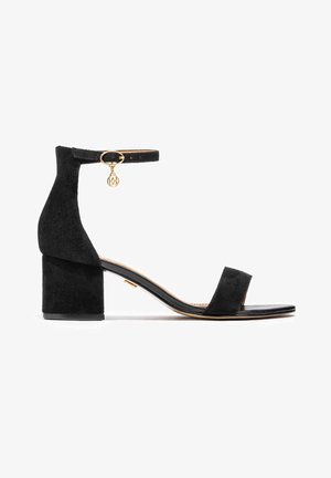 Black suede block-heel sandal with ankle strap, gold hardware accent, open toe, and flat sole. Features a minimalistic design and smooth texture.