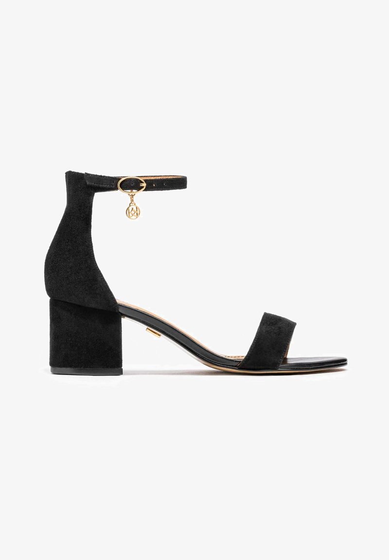 Black suede block-heel sandal with ankle strap, gold hardware accent, open toe, and flat sole. Features a minimalistic design and smooth texture.