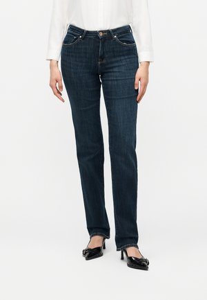 Kvinde iført mørkeblå jeans med lige ben, hvid langærmet skjorte og sorte spidse højhælede sko med spænde detaljer.