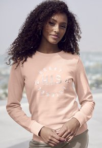 Sudadera de color rosa claro con texto blanco bordado en un diseño circular. Mangas largas con puños de canalé. La tela parece suave y texturizada.