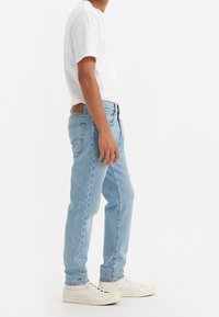 Ljusblå denimjeans med rak skärning, fem fickor och en brun läderlapp vid bakdelen av midjan, kombinerat med vita sneakers.