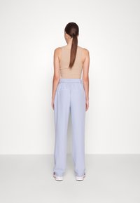 Kvinna med långt hästsvans, iförd en beige linne och ljusblå högmidjade byxor, står vänd mot en enkel vit bakgrund.