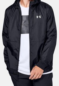 Chaqueta negra ligera con cremallera y capucha, puños elásticos y un logo en el pecho. Cuenta con una textura suave y bolsillos.
