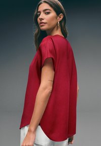 Red short-sleeved top with a relaxed fit, round neckline, and curved hem. Fabric appears smooth with a slight sheen.