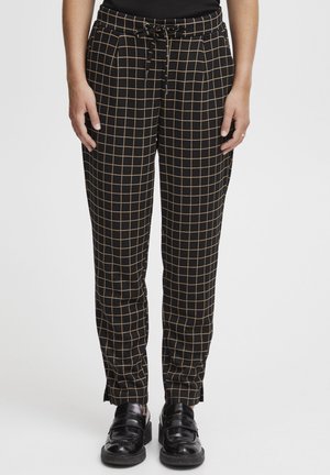Pantalons à carreaux noir et beige avec une taille élastique et des poches latérales ; jambe fuselée avec fentes aux chevilles. Portés avec des chaussures noires.