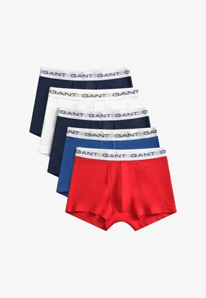 Homme torse nu aux cheveux courts et bouclés portant un boxer bleu marine avec une ceinture blanche marquée "GANT", debout devant un fond blanc uni.