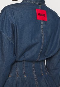 Abito in denim blu scuro dalla forma strutturata, con un'etichetta rettangolare rossa con la scritta "HUGO" cucita sul retro. Dettagli di cuciture fini.