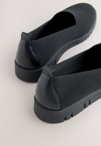 Chaussures noires slip-on avec dessus en maille texturée, languettes de talon en cuir et semelles épaisses en caoutchouc, vues de l'arrière en angle.