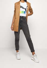 Ljusbrun blazer över en vit t-shirt med en svart Puma-logotyp och färgstark text, kombinerad med mörka högmidjade jeans och vita sneakers.