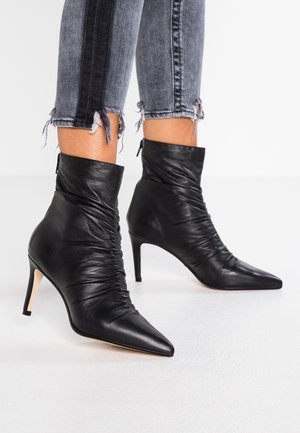 Černé kožené kotníkové boty s řasením, špičatými špičkami a jehlovými podpatky, nošené s roztrhanými šedými zkrácenými džíny.