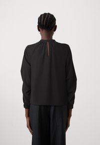 Blusa negra de manga larga con escote recogido y detalle de ojo de cerradura en la parte trasera. Confeccionada en tela suave, presenta puños elásticos y un corte relajado.