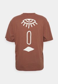 Brun bomulds T-shirt med et stort hvidt øje og geometriske figurer trykt på ryggen. Korte ærmer med rund halsudskæring.
