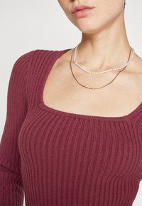 Ribbad, vinröd långärmad topp med fyrkantig halsringning, lager på lager med två halsband: en guldkedja och en pärlhalsband.