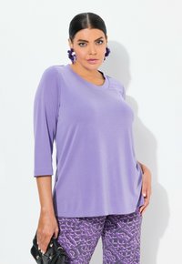 Lavendelfarbenes Oberteil mit drei Viertelärmeln aus weichem Stoff, kombiniert mit gemusterten lila Hosen. Das Oberteil hat einen runden Ausschnitt und einen lässigen Schnitt.