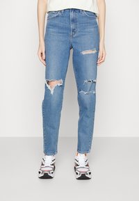Blue denim jeans with a high-waisted fit, featuring multiple large rips and frayed edges. Paired with gray and white sport shoes.