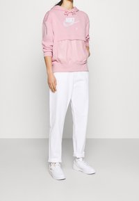 Rosa Nike hoodie med en framficka i kängurustil, vit logotext, i kombination med vita byxor och vita sneakers, designad för avslappnad användning.
