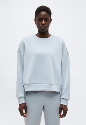 Žena s krátkymi vlasmi oblečená v svetlomodrej oversize mikine a zladených nohaviciach, stojaca pred jednoduchým bielym pozadím.